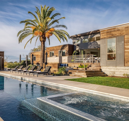 Sun-drenched modern home with wood-clad exterior, a sparkling lap pool, loungers, and a towering palm against a clear blue sky.