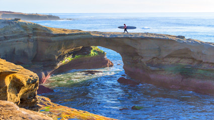 A vibrant San Diego coastal scene featuring a natural rock arch over the ocean, with a surfer walking atop the arch. The blue waters and bright sky evoke the fresh, active lifestyle and year-round appeal of warm weather destinations.
