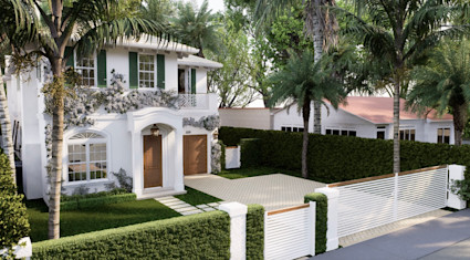 Lush tropical streetscape view of a white two-story home with green shutters, herringbone driveway, and palm canopy.