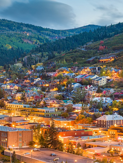 An elevated twilight view of Park City, Utah, with glowing golden lights across a hillside town nestled in lush, pine-covered mountain ridges.