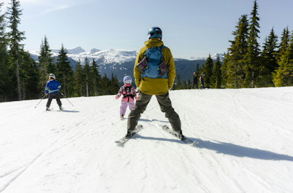 A man and two children skiing down a slope surrounded by pine trees.