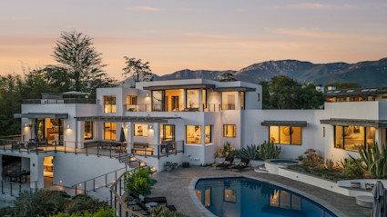 Modern multilevel white stucco estate at twilight with warm interior lighting, pool, and Santa Barbara mountain views.