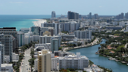 A panoramic view of Miami, Florida, featuring high-rise buildings, a vibrant blue ocean, and a winding waterway lined with palm trees. The cityscape is bustling with life, highlighting Miami's appeal as a luxurious warm-weather destination for snowbirds seeking sunshine during the winter season.