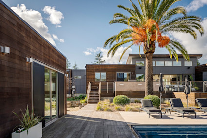 Modern wood-clad home with palm tree, pool, and lounge chairs under a blue sky.