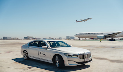 A PS branded BMW car park on the tarmac with a Japan Airlines plane behind it and another plane in the air right after take off.
