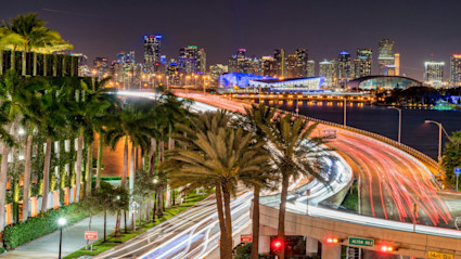 A vibrant night scene of Miami, Florida, featuring light trails from vehicles, palm trees, and the illuminated city skyline. The lively atmosphere and glowing buildings capture the essence of Miami's energetic nightlife and urban luxury.