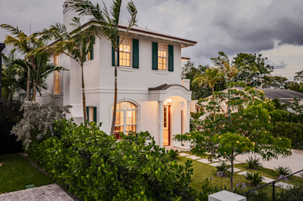 White two-story coastal home with green shutters and warm-lit windows at dusk, framed by palms and tropical greenery.