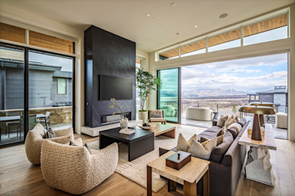 Modern living room with floor-to-ceiling windows, stone fireplace, and sweeping mountain views opening to a furnished outdoor terrace.