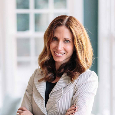 Pe imagine este reprezentată o femeie cu părul roșcat , imbractă într-un sacou alb și tricou gri. Femeia zâmbește