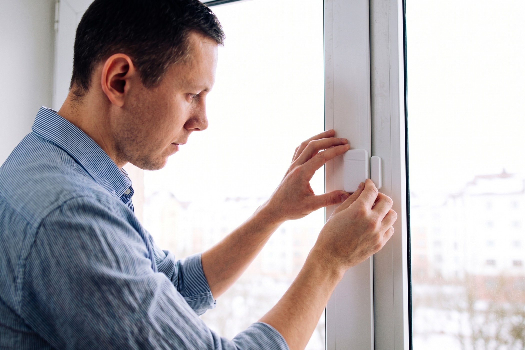 A man installing a white smart window sensor on the frame of a glass window.