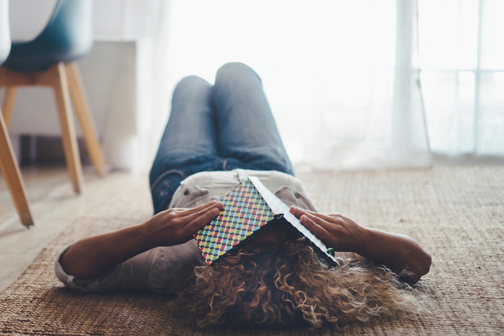 Een studerende vrouw met stress ligt op de grond in haar woning en houd een opengeslagen studieboek voor haar gezicht.