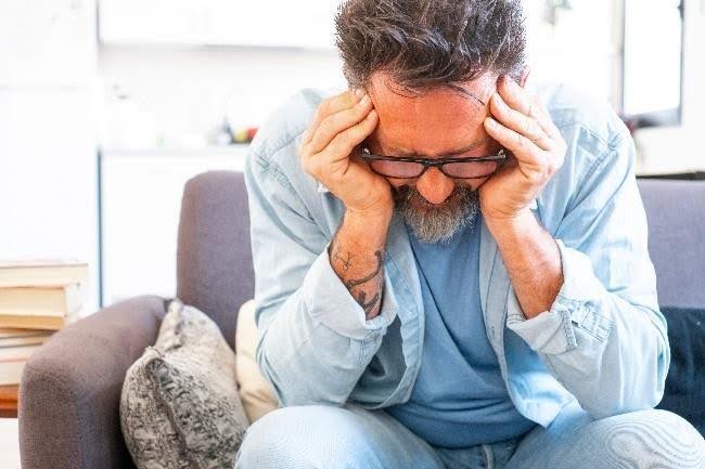 Man zit op de bank, kijkt naar beneden en drukt zijn vingertoppen tegen zijn voorhoofd aan