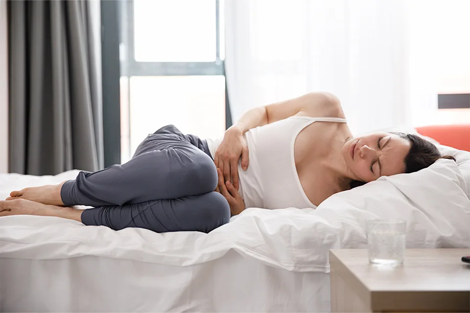 La femme est allongée sur le lit, les jambes repliées. Grimaçant de souffrance, elle se tient le ventre à deux mains.