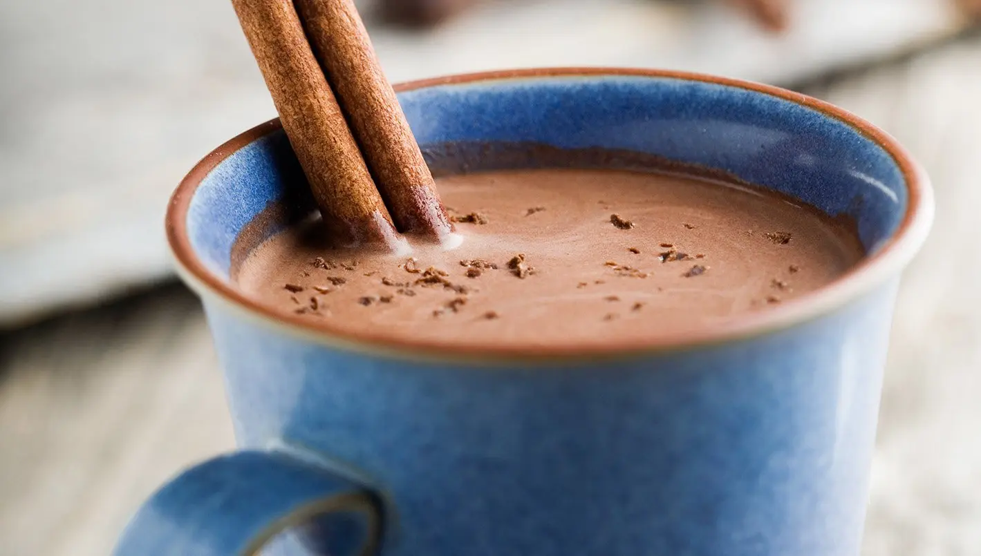 Rich hot chocolate in a cozy mug, topped with whipped cream and chocolate drizzle, set on a wooden table with a warm blanket and candles for a calming evening ambiance