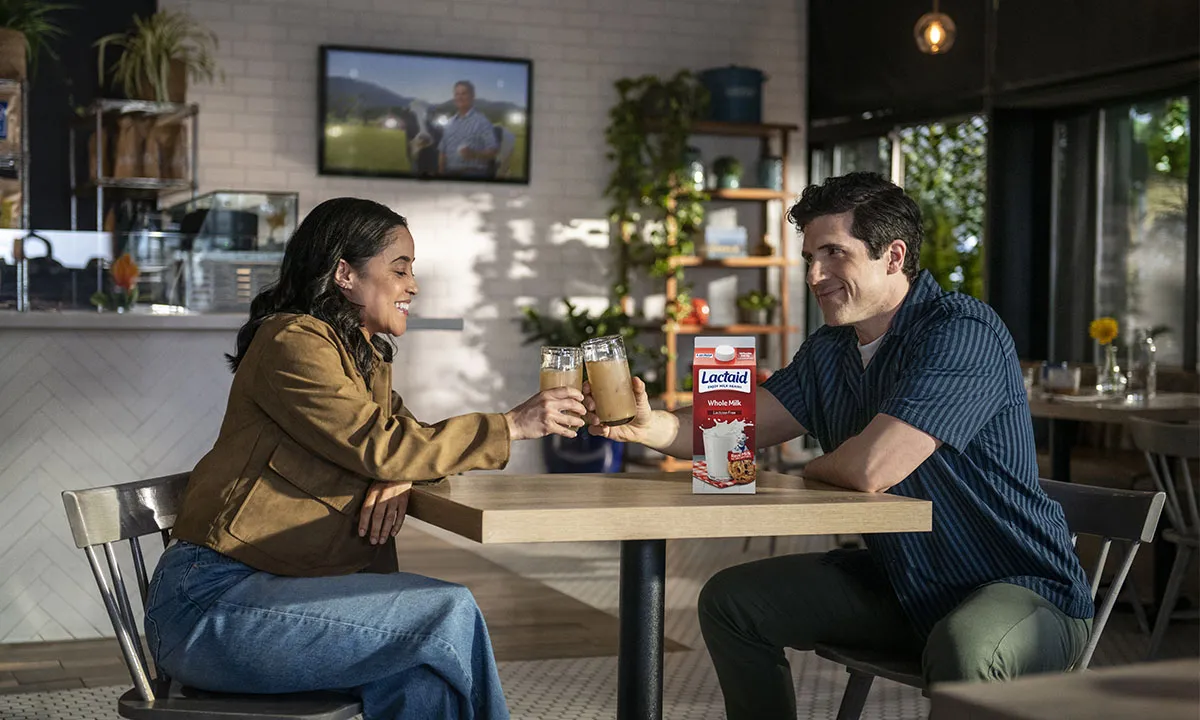 Two people toasting with glasses of iced latte at a café table, with a carton of Lactaid Whole Milk displayed.