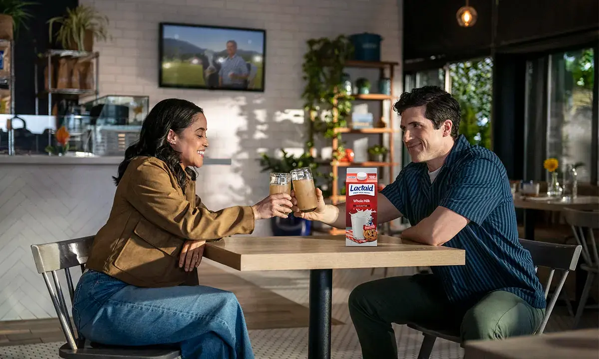 Two people toasting with glasses of iced latte at a café table, with a carton of Lactaid Whole Milk displayed.