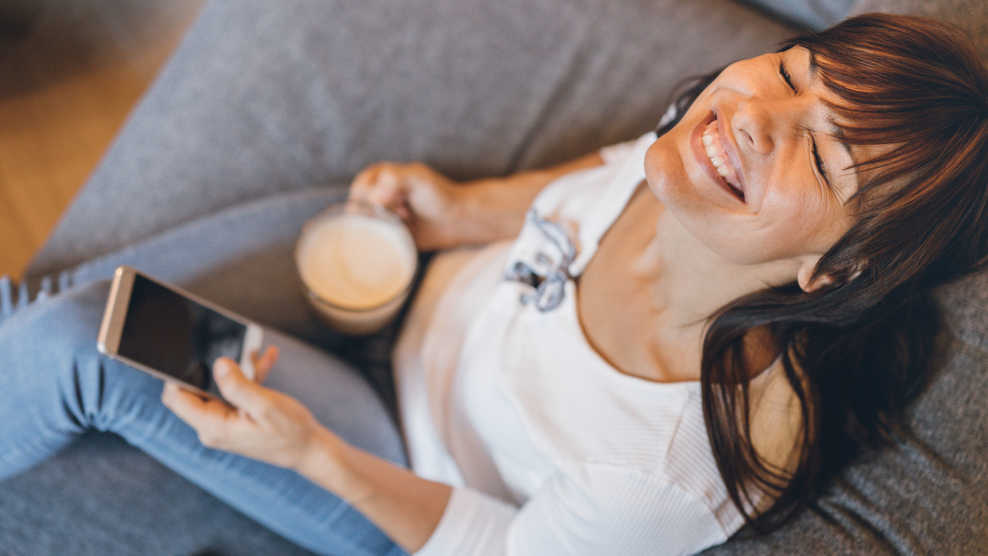 Frau sitzt glücklich mit einer Tasse Kaffee auf der Couch und verwendet dabei ihr Handy