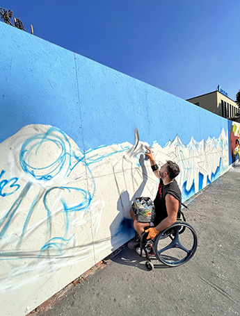 L’artista Kasy23 lavora alla sua opera in sedia a rotelle, condizione che l’ha spinto ad innovare ulteriormente la sua arte.
