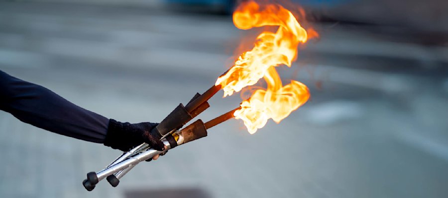 Une main gantée tenant deux torches enflammées avec des flammes orange vif contre un fond flou gris-bleu.