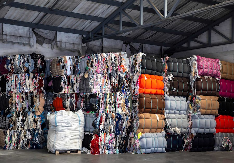 Grote stapels gecomprimeerd textielafval in verschillende kleuren, gebundeld in balen in een recyclingloods met metalen dak.
