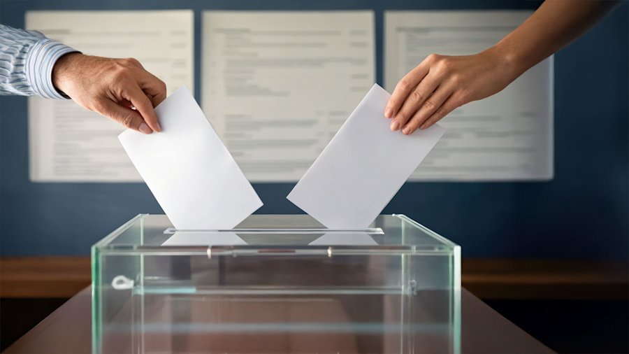 Twee handen stoppen stembiljetten in een transparante stembus tijdens een verkiezing, met kieslijsten op de achtergrond.
