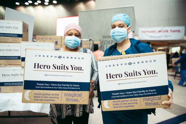 Two health care workers in ppe stand in front of a stack of dinnerly meal kits and hold their own boxed meal kits. 