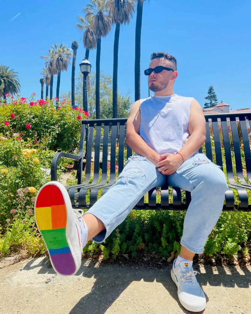 Creator poses on bench wearing Sperry PFLAG rainbow bottom shoes