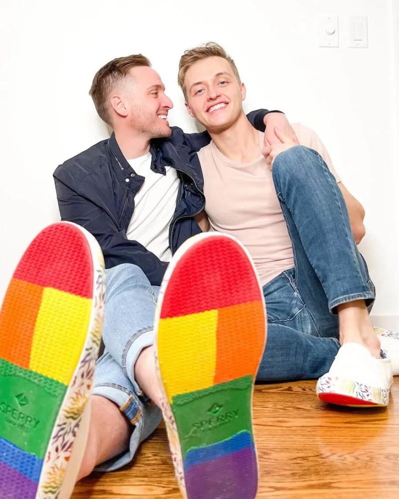 A couple poses sitting against wall in Sperry PFLAG rainbow bottom shoes