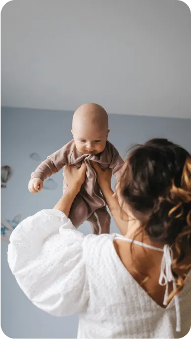Woman in white dress lifts baby in brown onesie in the air