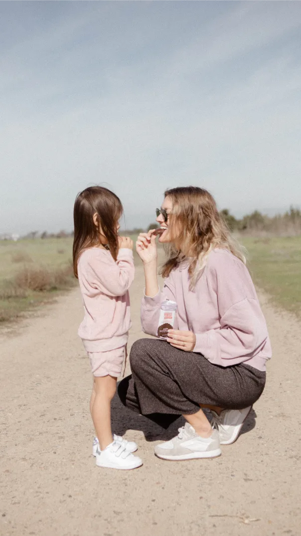 Woman crouches down in a dirt path to share a packaged snack bar with a dark haired child