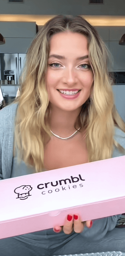 Creator with blonde hair smiles at the camera while holding a closed box of Crumble cookies