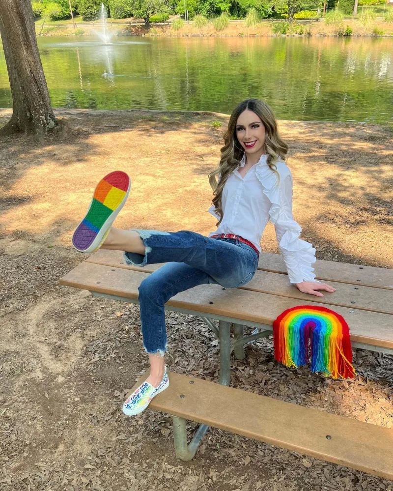 Creator leans on bench by a lake wearing Sperry PFLAG rainbow shoes