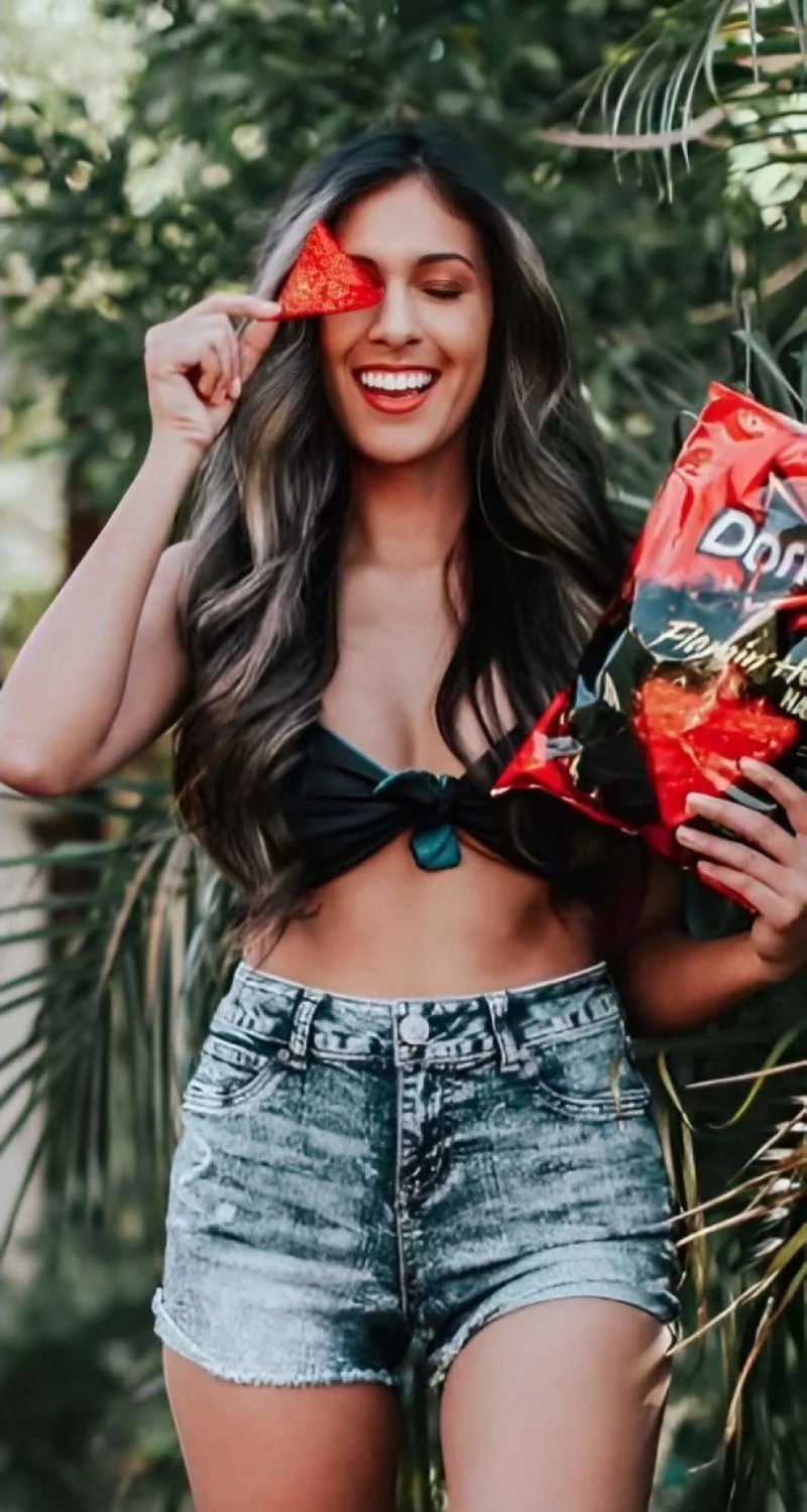 Woman influencer in tropical location holding Dorito over left eye and bag of Doritos Flamin Hot Nachos in right hand