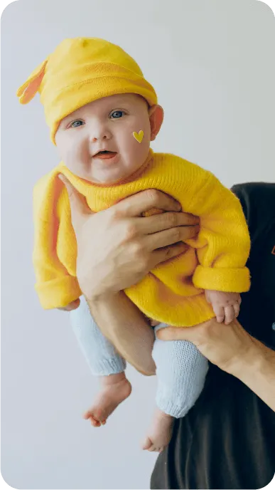 Baby held to smile at camera wears yellow sweater and matching yellow hat