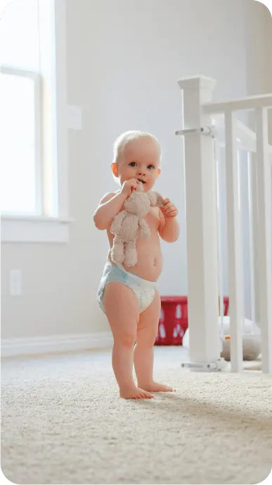 Baby wearing diaper chews on bunny toy while standing in nursery