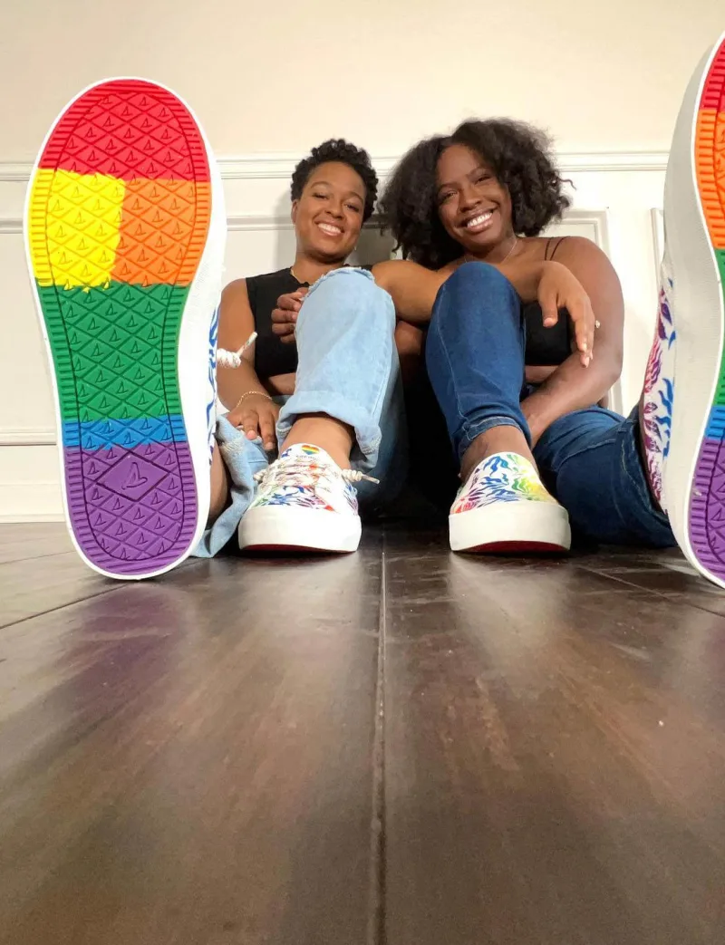 Two women lean against wall smiling wearing Sperry PFLAG shoes