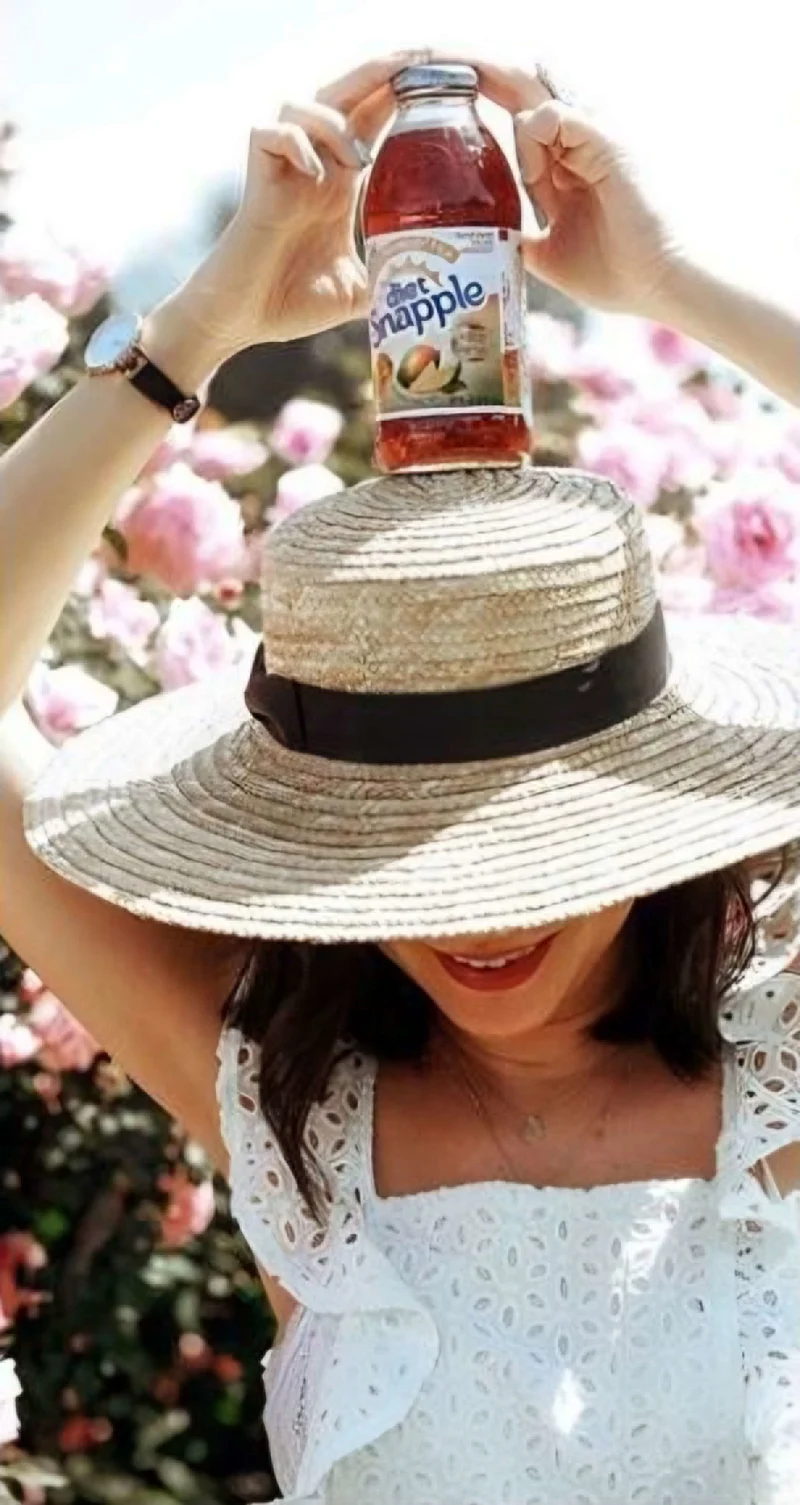 Influencer in sun hat looking down surrounded by pink flowers on a sunny day holding a diet Snapple drink on top of her head