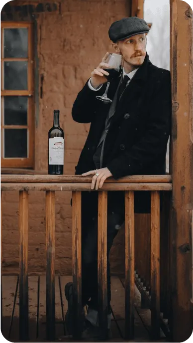 Man in cap drinks a glass of red wine on a covered porch in the snow