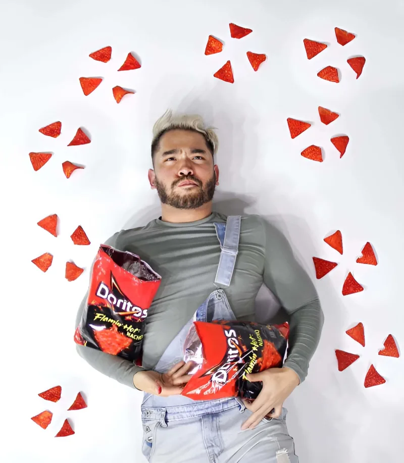 Man influencer eating Doritos lying down on floor scattered with Doritos holding two bags of Doritos Flamin Hot Nachos