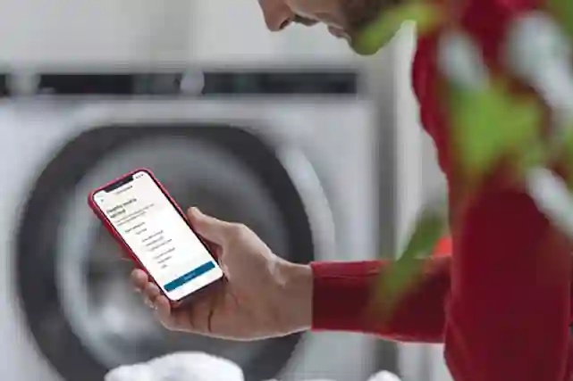 Henkilö katsoo puhelimesta taustalla näkyvän pyykkikoneensa tietoja.