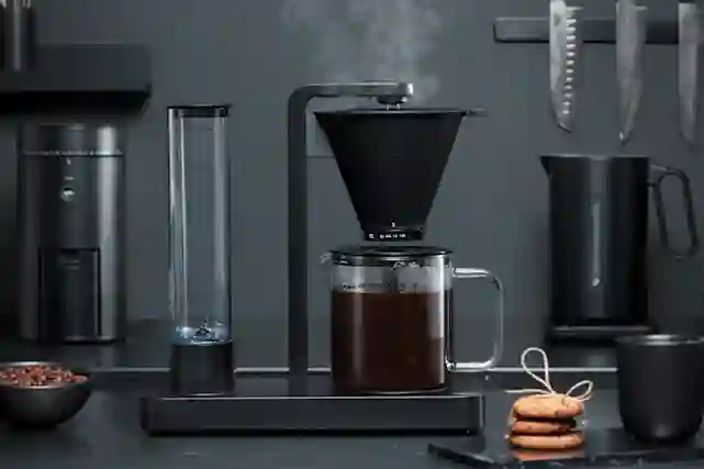 Harmaassa keittiössä Wilfan mustia pienkoneita. Musta kahvinkeitin keskiössä.