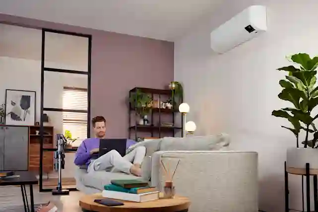 Mies hymyilee sohvalla läppäri sylissä ilmalämpöpumpun alla, verkkarit jalassa.