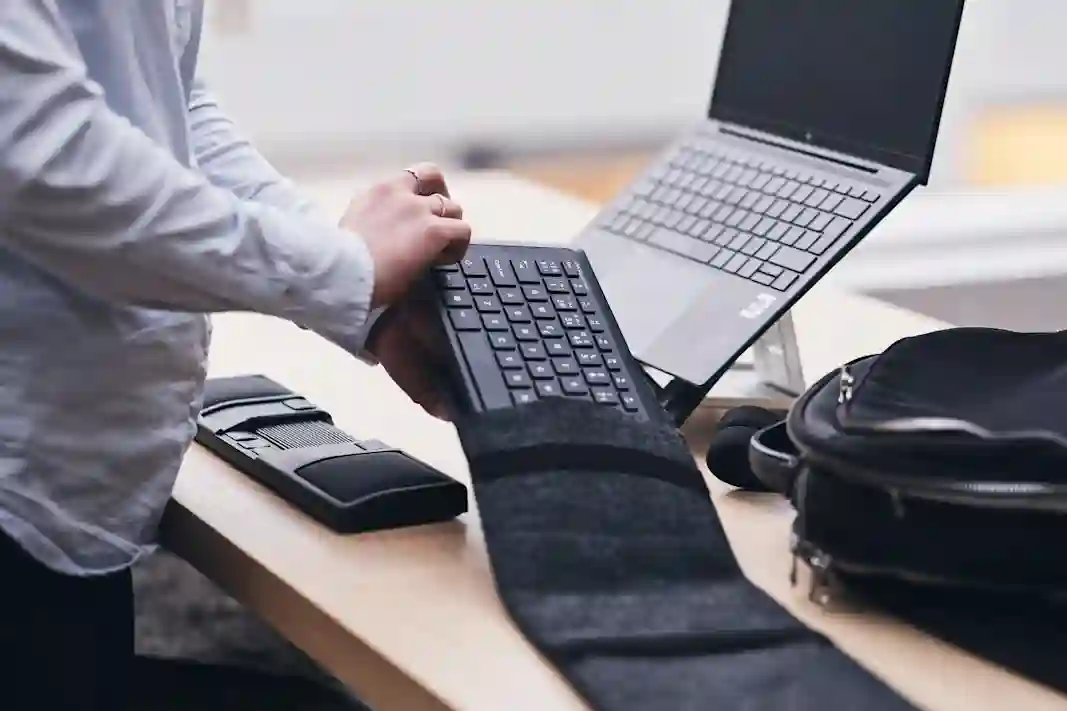 A person taking a compact keyboard out of a bag at a desk with an open laptop and work tools.