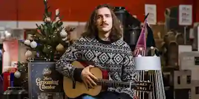 A man in a Christmas sweater with a guitar, next to a Men's Grooming Calendar package and a decorated Christmas tree.