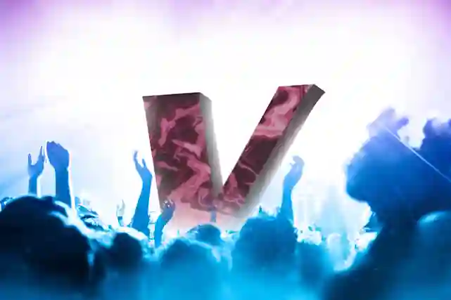 Esiintymislavalla viininpunainen V-logo näyttävien valojen ympäröimänä. Kuva otettu yleisön joukosta, ja juhlivia katsojia näkyy lavan edustalla.