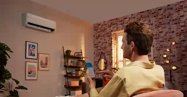 Mies istuu nojatuolilla puhelin kädessä, jossa auki ilmalämpöpumpun tiedot. Seinällä ilmalämpöpumppu.