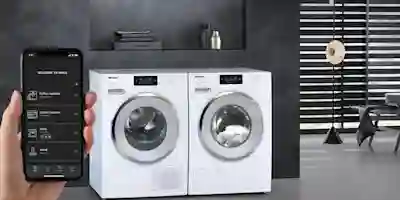 A hand holds a smartphone with the Miele app, with two white Miele tumble dryers in the background in a gray laundry room.