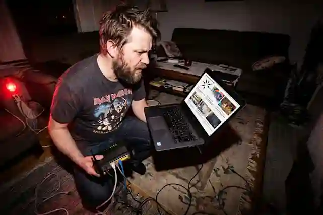 Mies kärvistelee olohuoneessa reitittimen kanssa. Hänellä on läppäri pöydällä ja lukee Verkkokauppa.comin opasta asiasta.