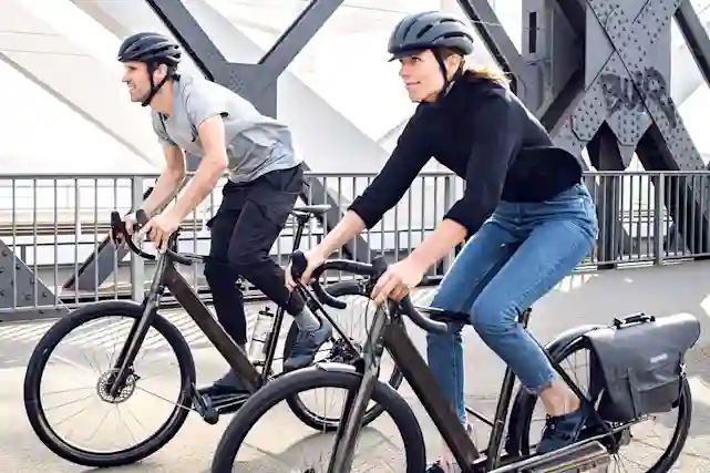 A man and woman ride electric bikes wearing helmets near a modern bridge in an urban setting.