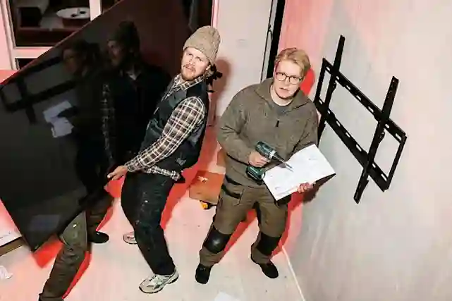 Kolme kaverusta yrittää asentaa televisiota seinälle. Kaksi kantaa televisiota ja yksi yrittää lukea ohjeita pora kädessään. Lue tämä opas niin tiedät tästä projektista enemmän.